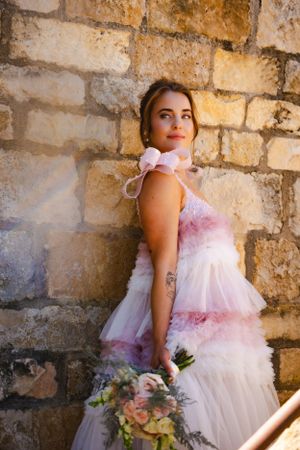 A romantic bridal portrait against a stone wall with a flowing pink and white tulle dress and delicate floral bouquet.