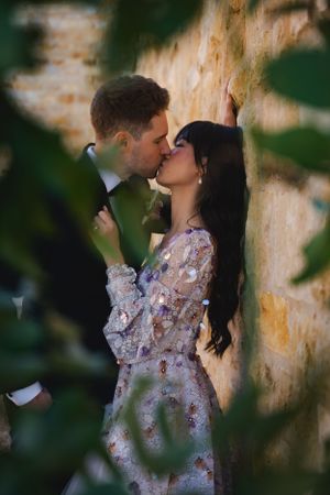 A romantic kiss shared between bride and groom against a rustic wall with green foliage in the foreground at the Sunstone Winery.