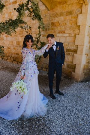 A couple in wedding attire poses against a stone wall with climbing vines, creating a romantic Mediterranean atmosphere.