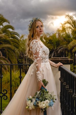 Bride in lace long sleeve wedding dress holds floral bouquet with blue and white flowers on balcony at sunset during a destination wedding.