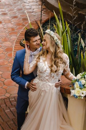 A romantic wedding couple embraces by a brick wall with tropical plants and floral decorations.