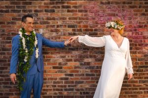 A couple in wedding attire holding hands against a brick wall background, adorned with flower garlands.