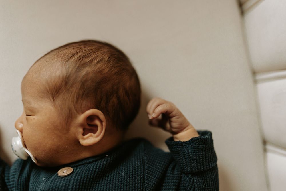 Sleeping newborn baby wearing dark sweater on white cushion.