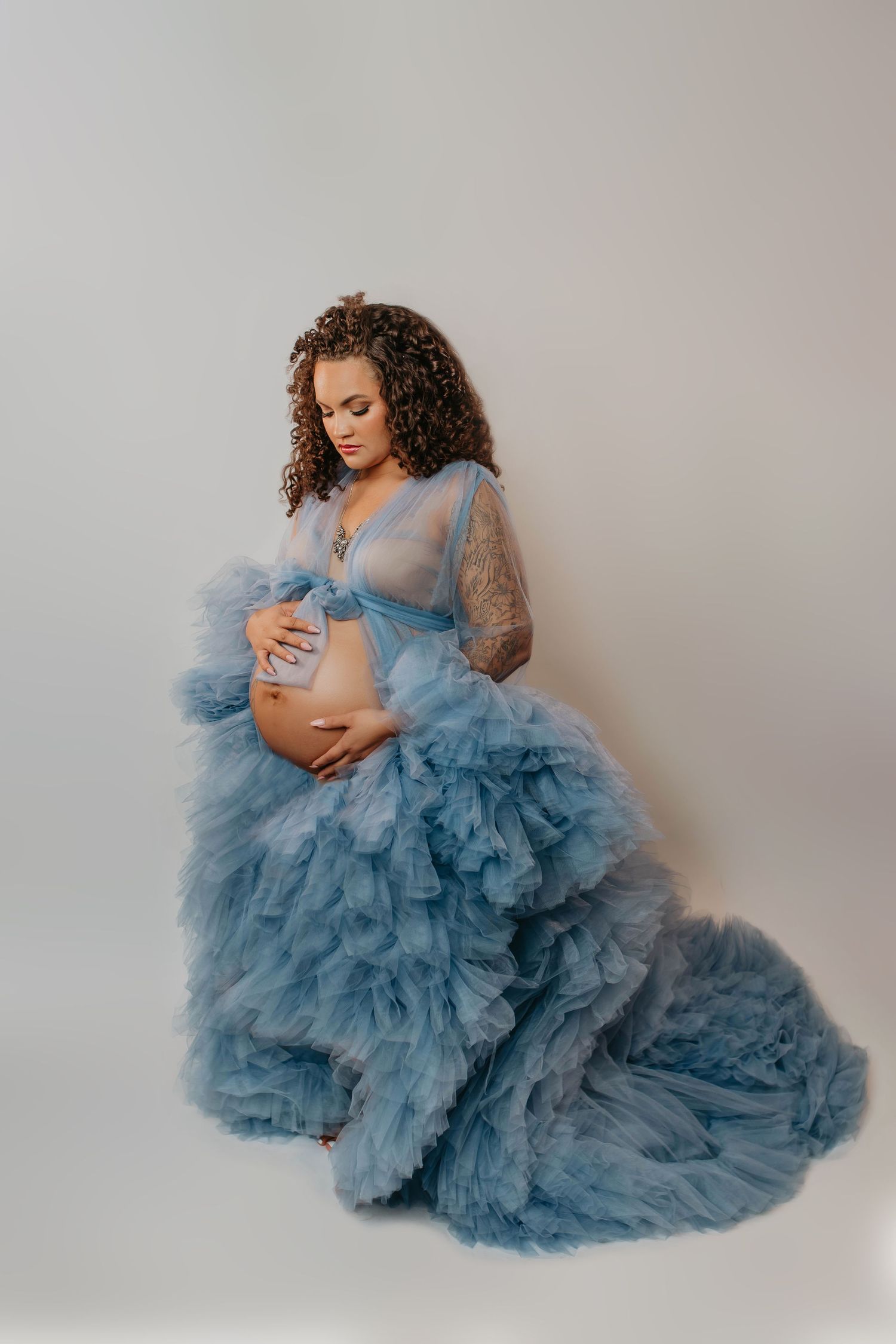 Maternity portrait of woman in flowing blue tulle gown against light background.