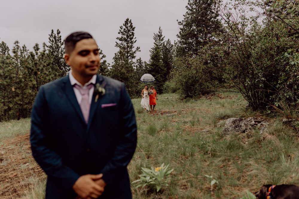 A groom in a navy suit stands in an outdoor wooded setting while a figure in red appears in the background.