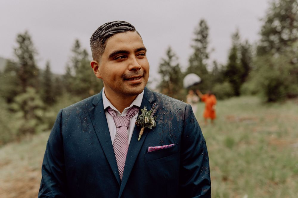 A groom in a navy suit and pink shirt smiles while wearing a boutonniere in an outdoor setting.