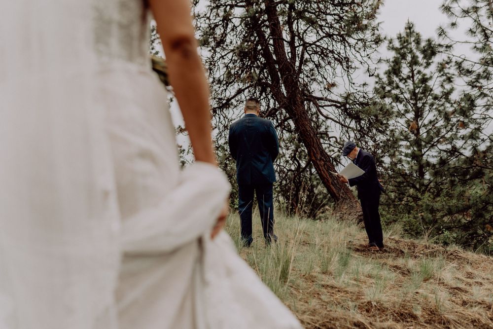 Wedding dress detail in foreground with ceremony participants visible on hillside.