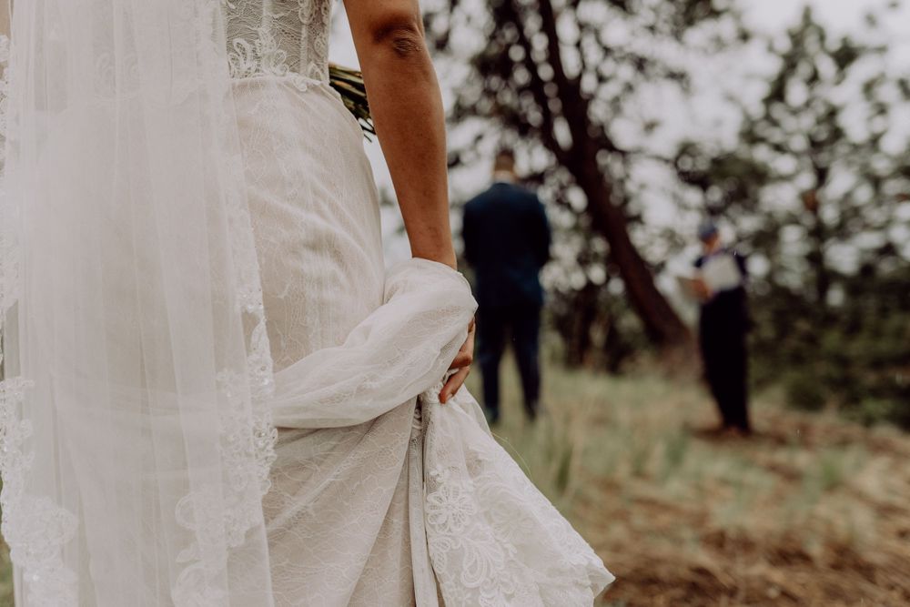 View of wedding dress detail with suited figures visible in the background outdoors.