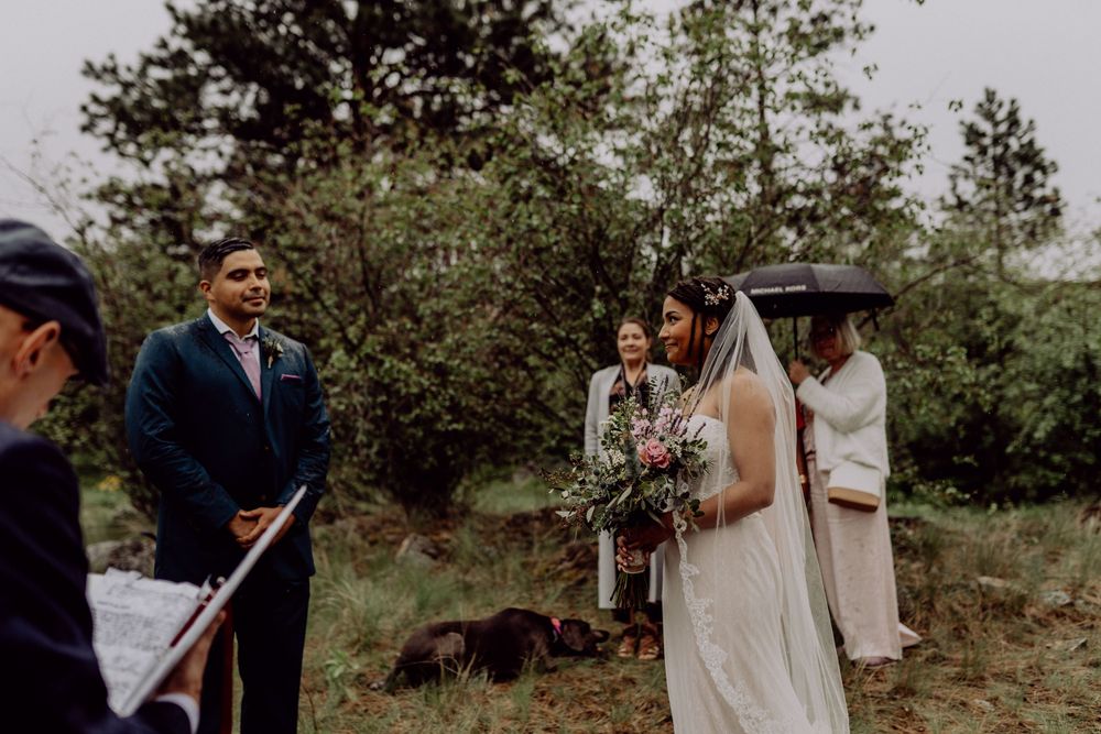 A moody outdoor wedding ceremony surrounded by pine trees with attendees holding umbrellas.