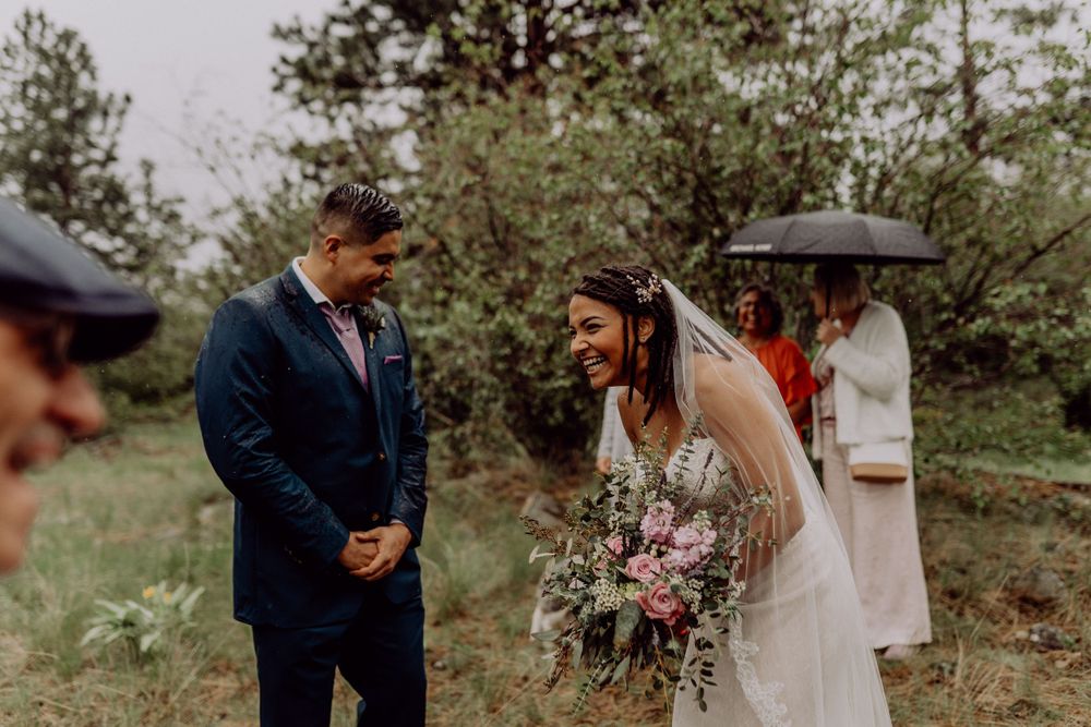 Outdoor wedding ceremony in a natural setting with pine trees and an officiant holding an umbrella.