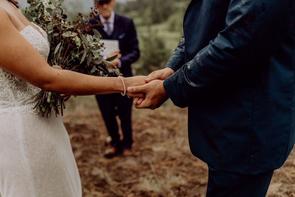 Close up of wedding couple holding hands during outdoor ceremony with bouquet.