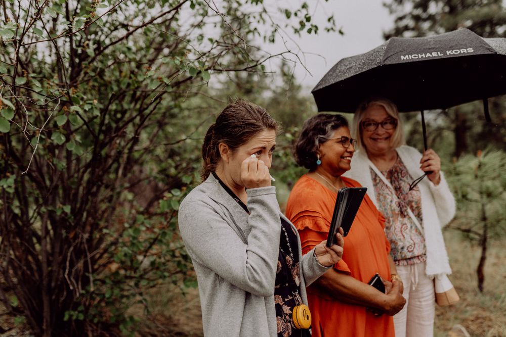 Wedding guests with umbrellas watching ceremony in wooded setting.