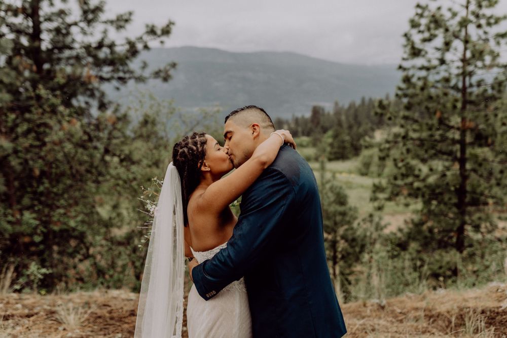 A romantic wedding kiss with mountain scenery and evergreen trees in the background.