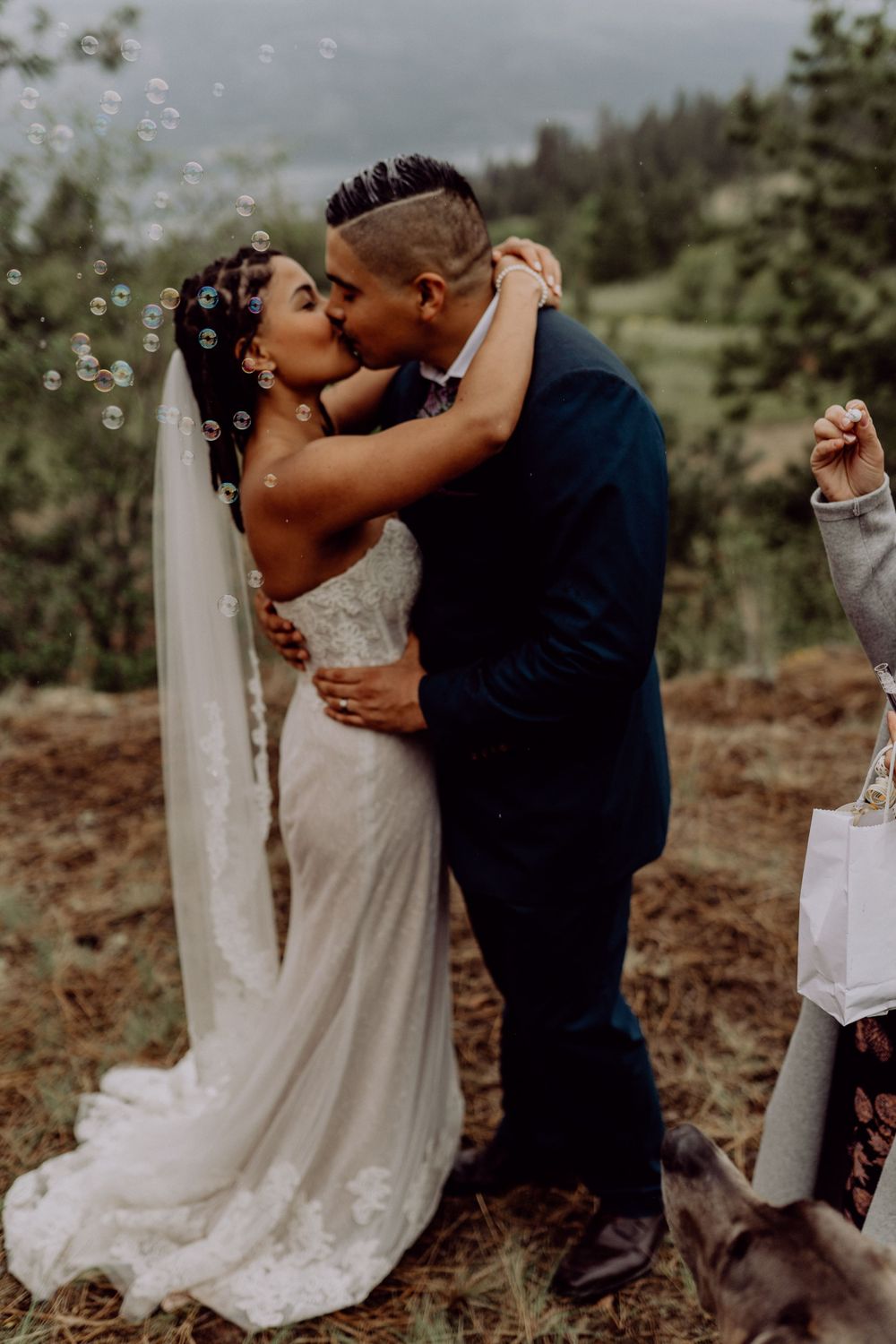 Newlyweds share their first kiss surrounded by bubbles in a mountain setting.