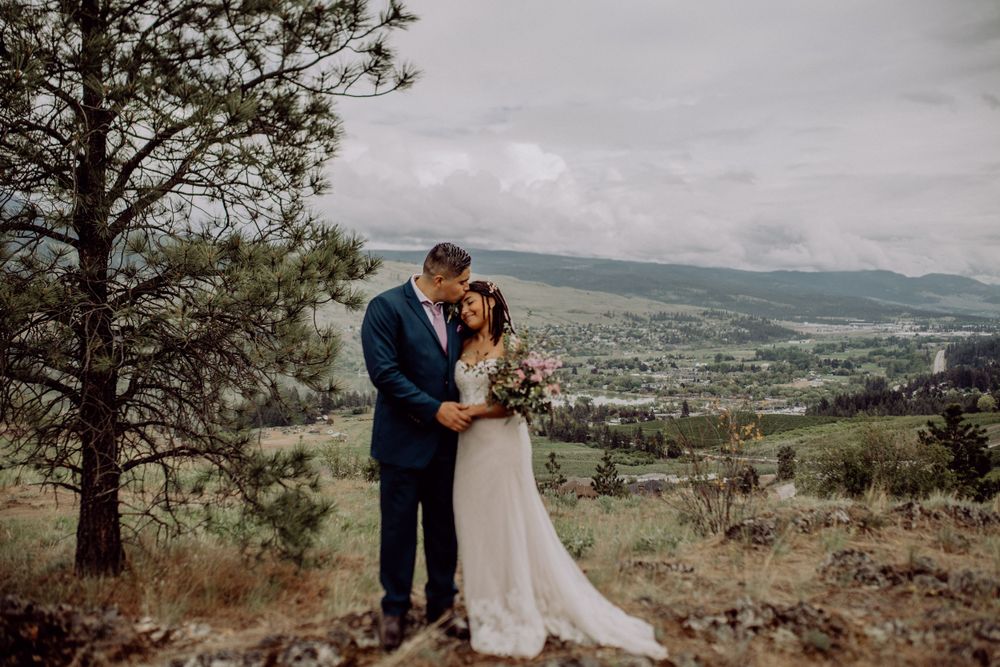 Wedding couple embraces on a hilltop overlooking a scenic mountain valley landscape.