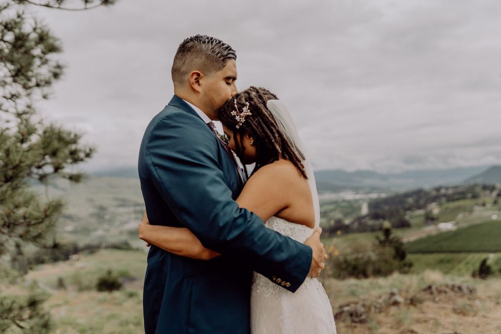 A romantic wedding moment captured on a scenic hilltop overlooking rolling valleys under cloudy skies.