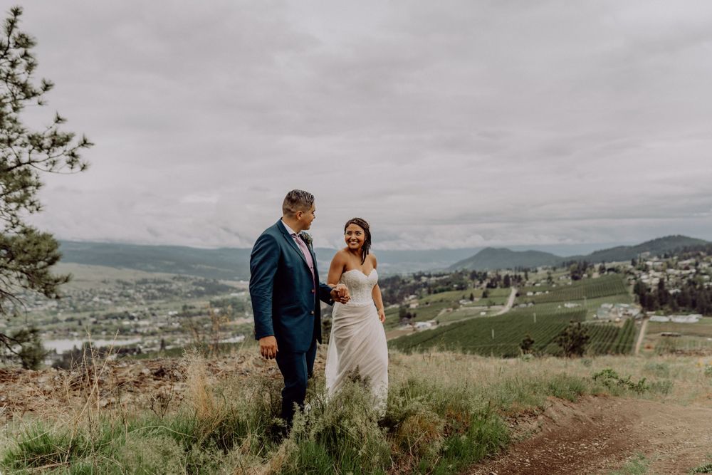 A wedding couple stands together on a hilltop meadow with panoramic mountain views in the background.