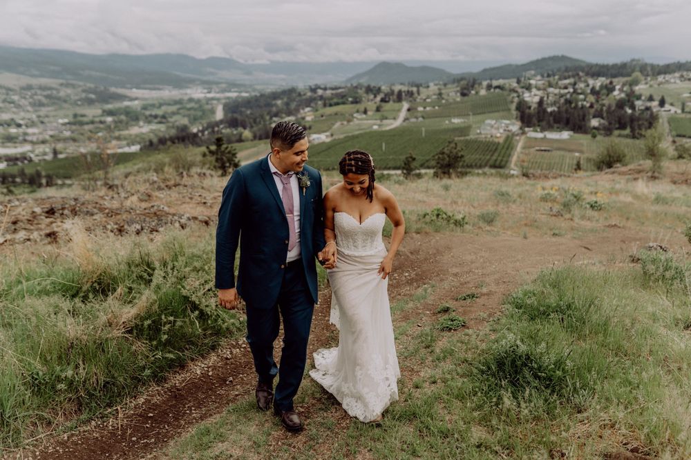 A couple in wedding attire walks along a dirt path on a mountainside with valley views.
