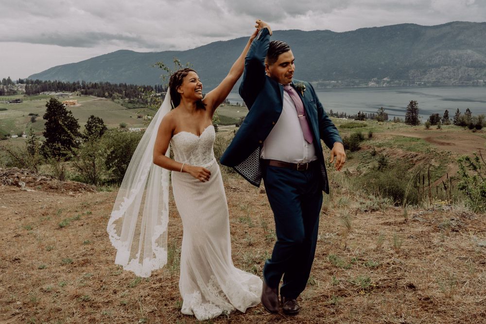 A joyful couple dances on a mountainside with a lake and mountains in the background.