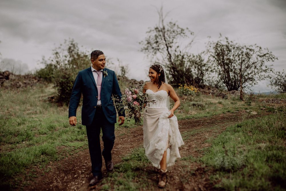 A couple walks hand in hand along a dirt path in nature wearing wedding attire on an overcast day.