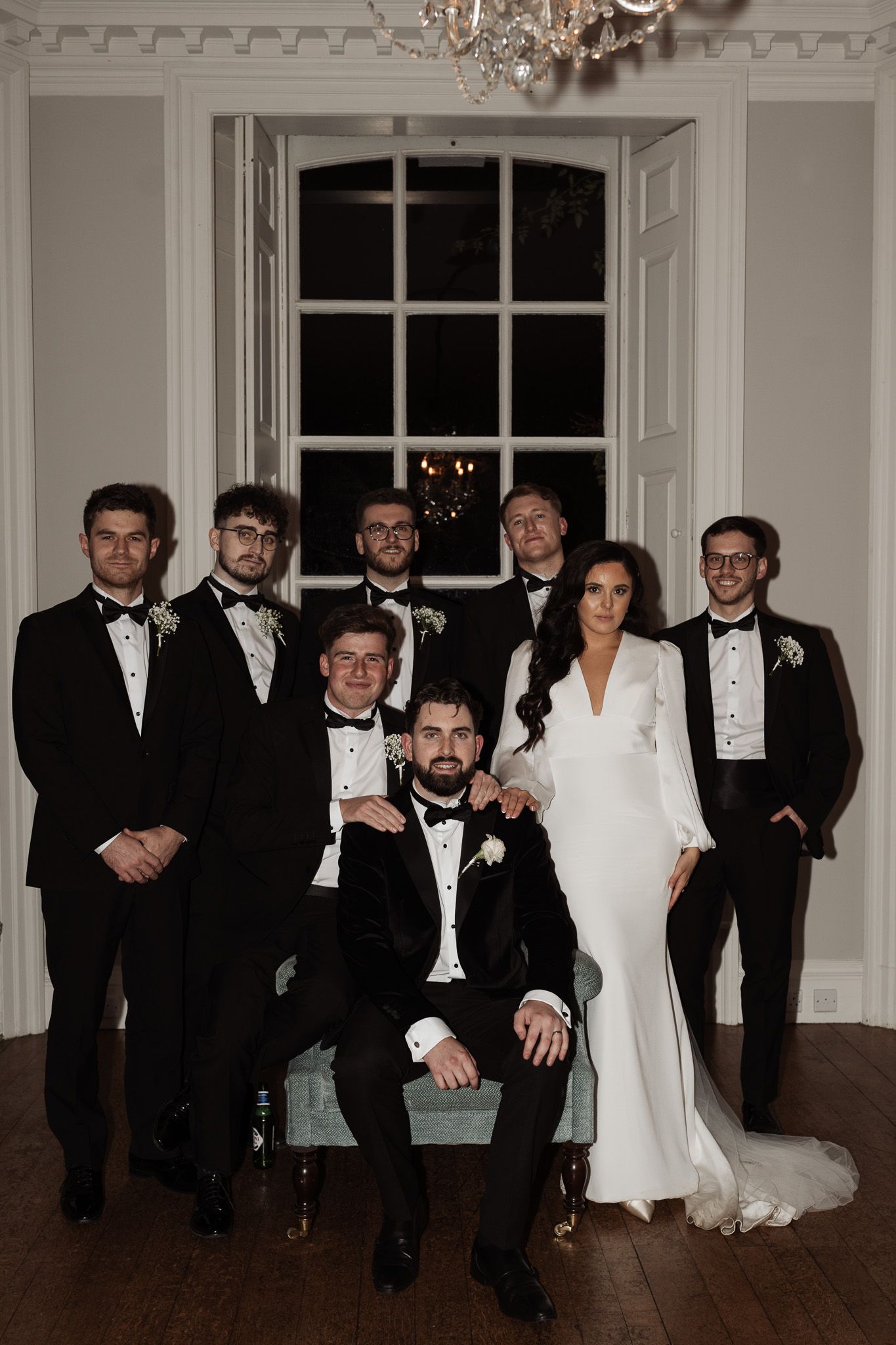 Formal wedding party portrait in a classic room with ornate molding and chandelier.