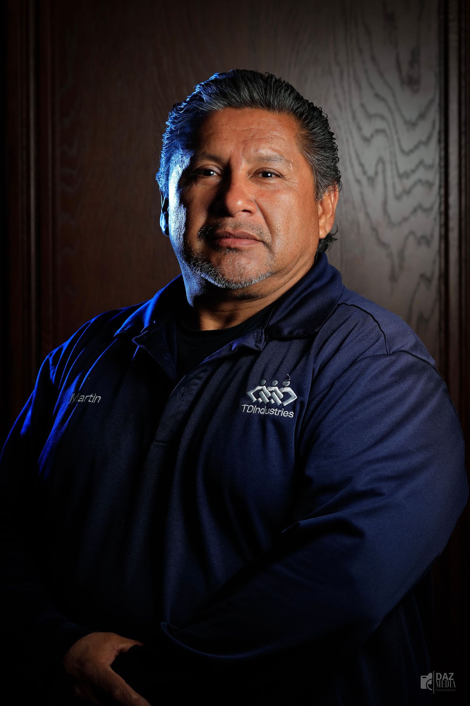 Professional headshot of person in navy blue uniform against dark wooden background with dramatic lighting.