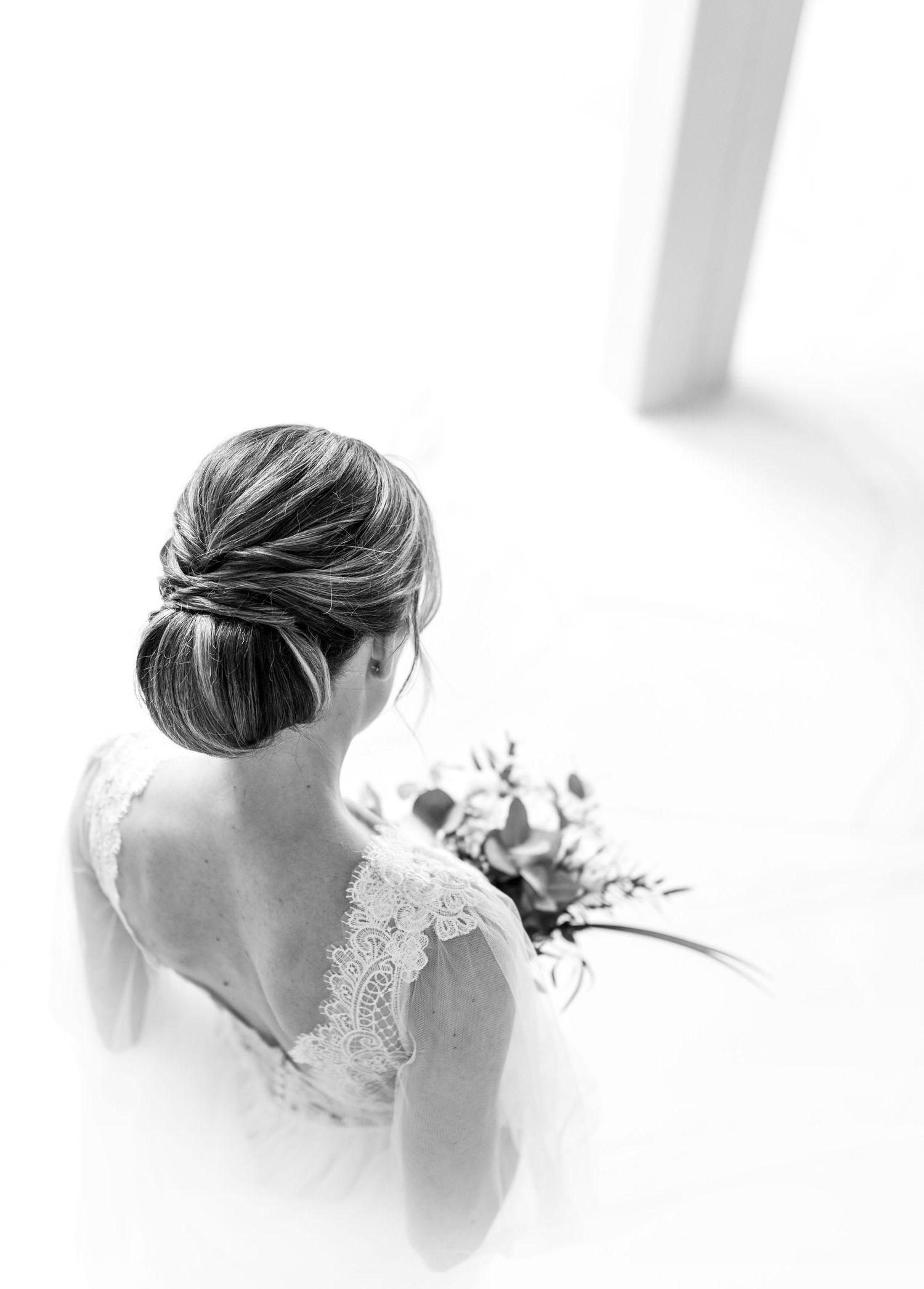 Elegant bride in lace dress shown from behind with updo hairstyle holding bouquet.