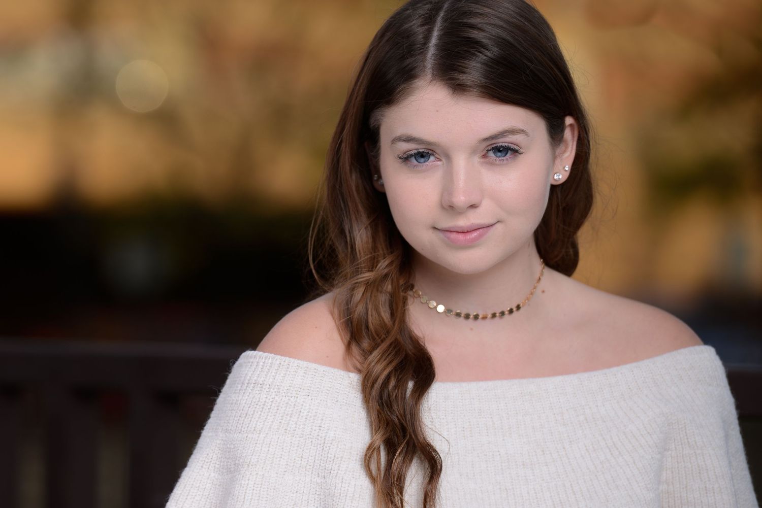 Closeup portrait wearing an off-shoulder white top and delicate necklace against a softly blurred background.