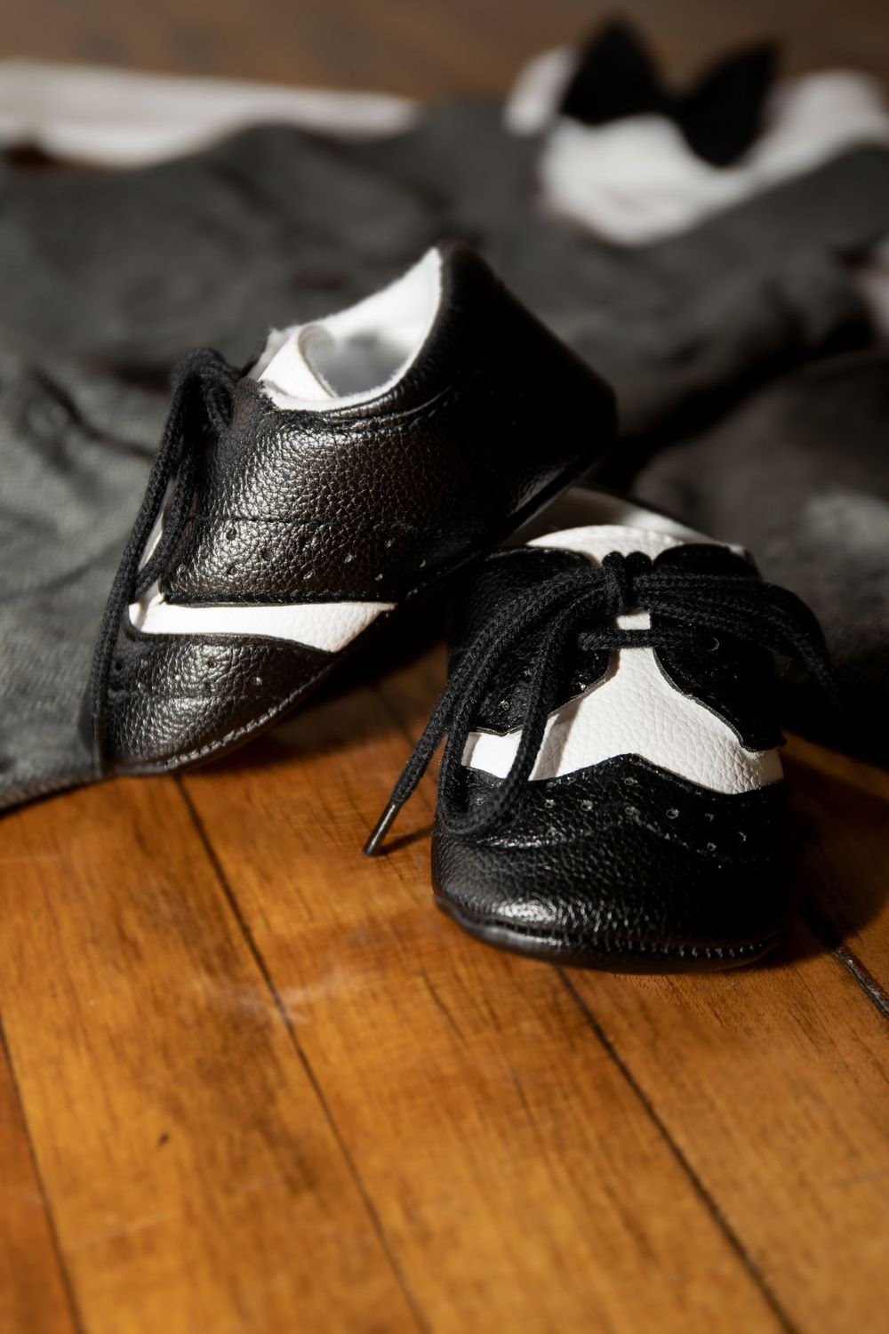 Black and white leather oxford baby shoes sit on a wooden floor.