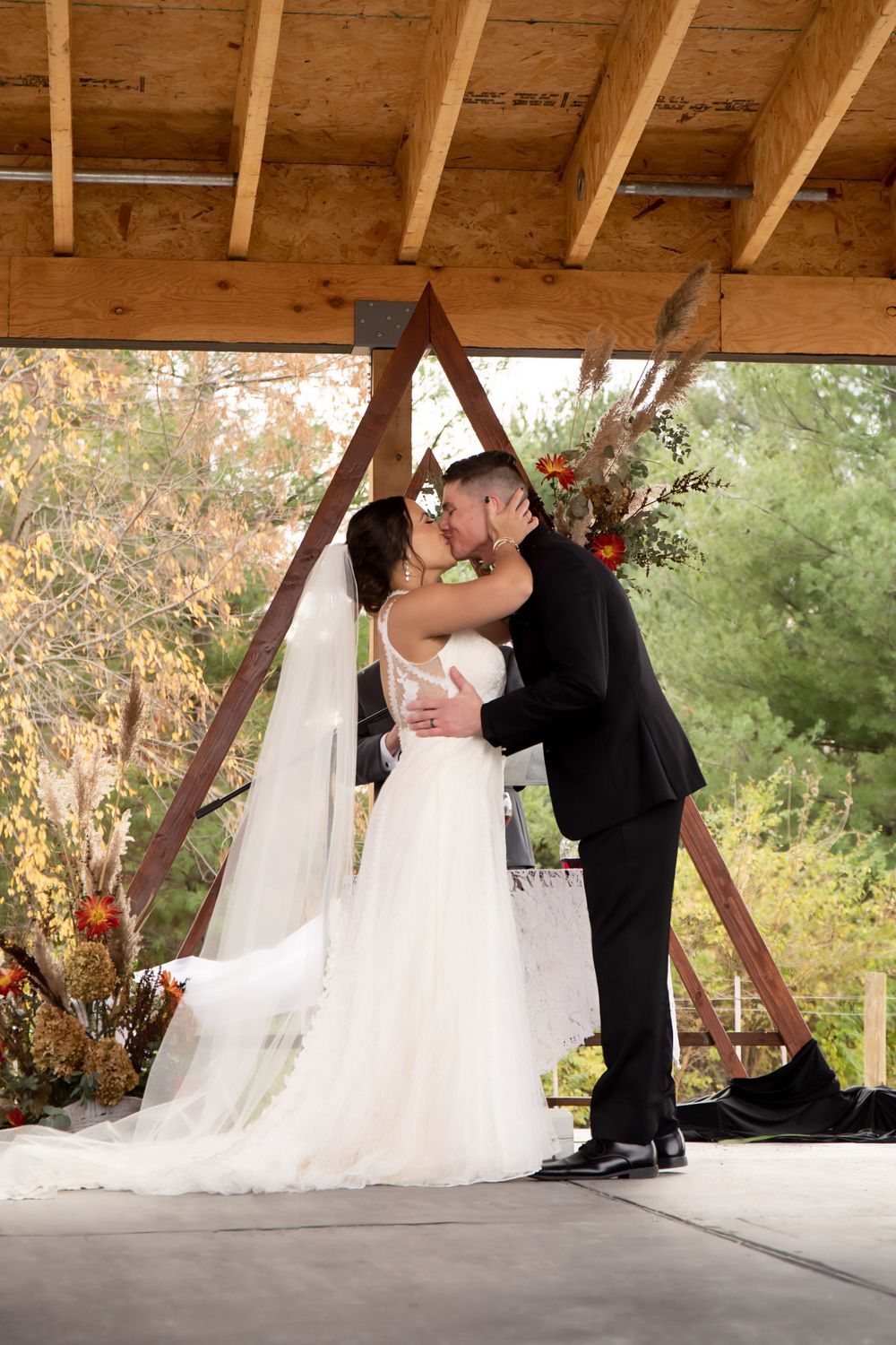 Wedding couple kisses under wooden triangular arch with autumn decor.