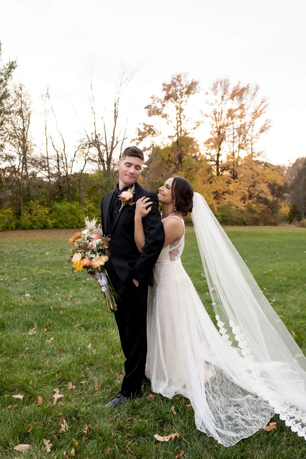 Newlyweds share a joyful moment outdoors in autumn with a flowing lace veil and colorful bouquet.