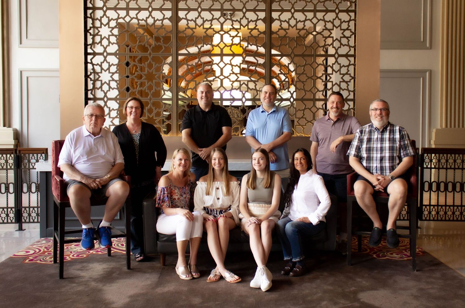 Group portrait of people seated on chairs against a decorative wall with geometric patterns.