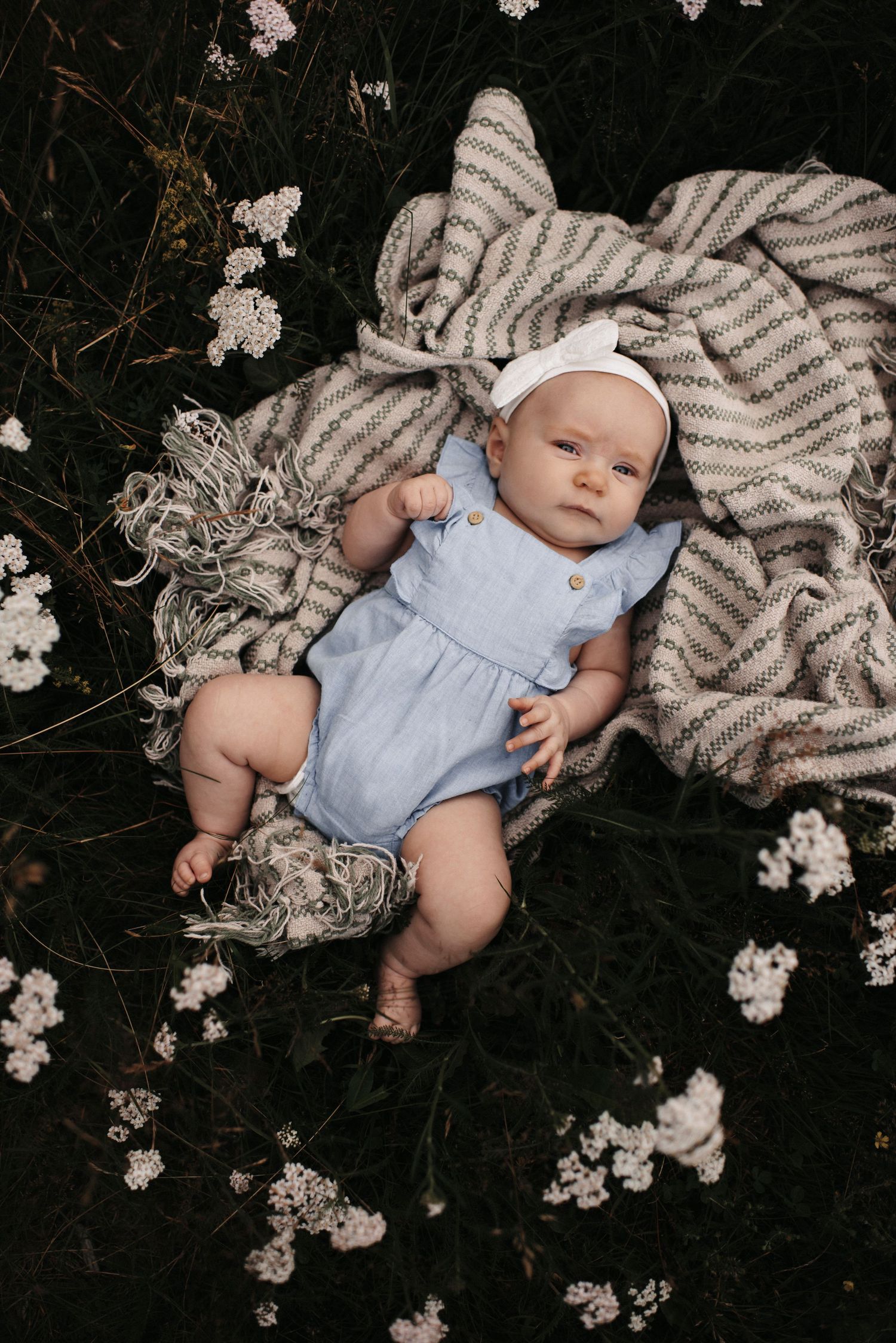Baby in light blue romper lying on striped blanket surrounded by white wildflowers.
