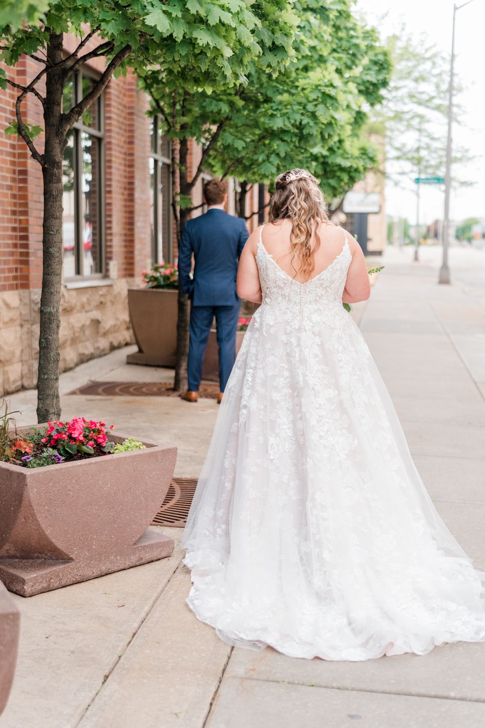 A bride in a flowing white gown walks along a city sidewalk lined with trees and planters.