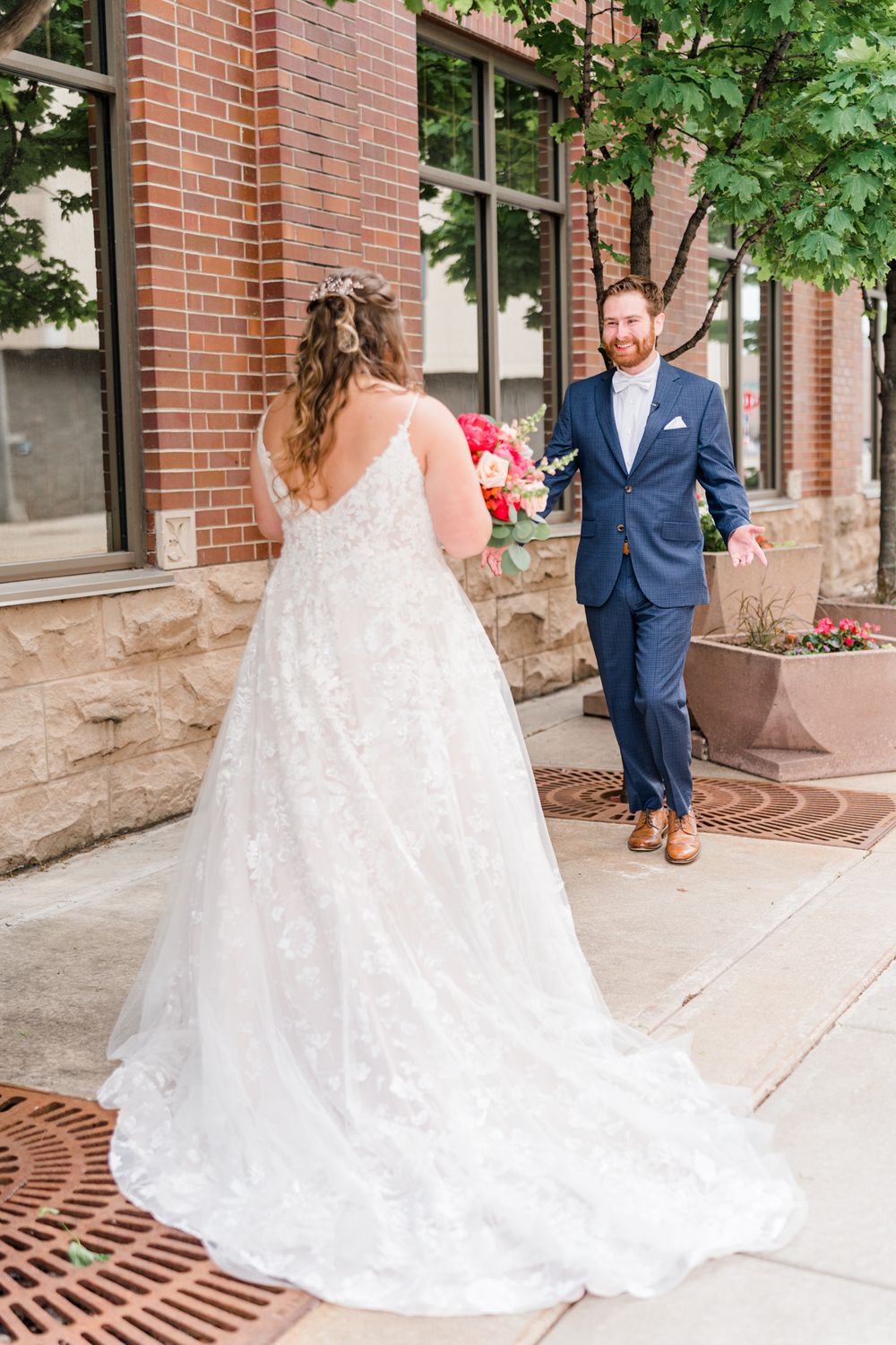 A bride in a flowing lace gown reveals herself to her groom on a city sidewalk.