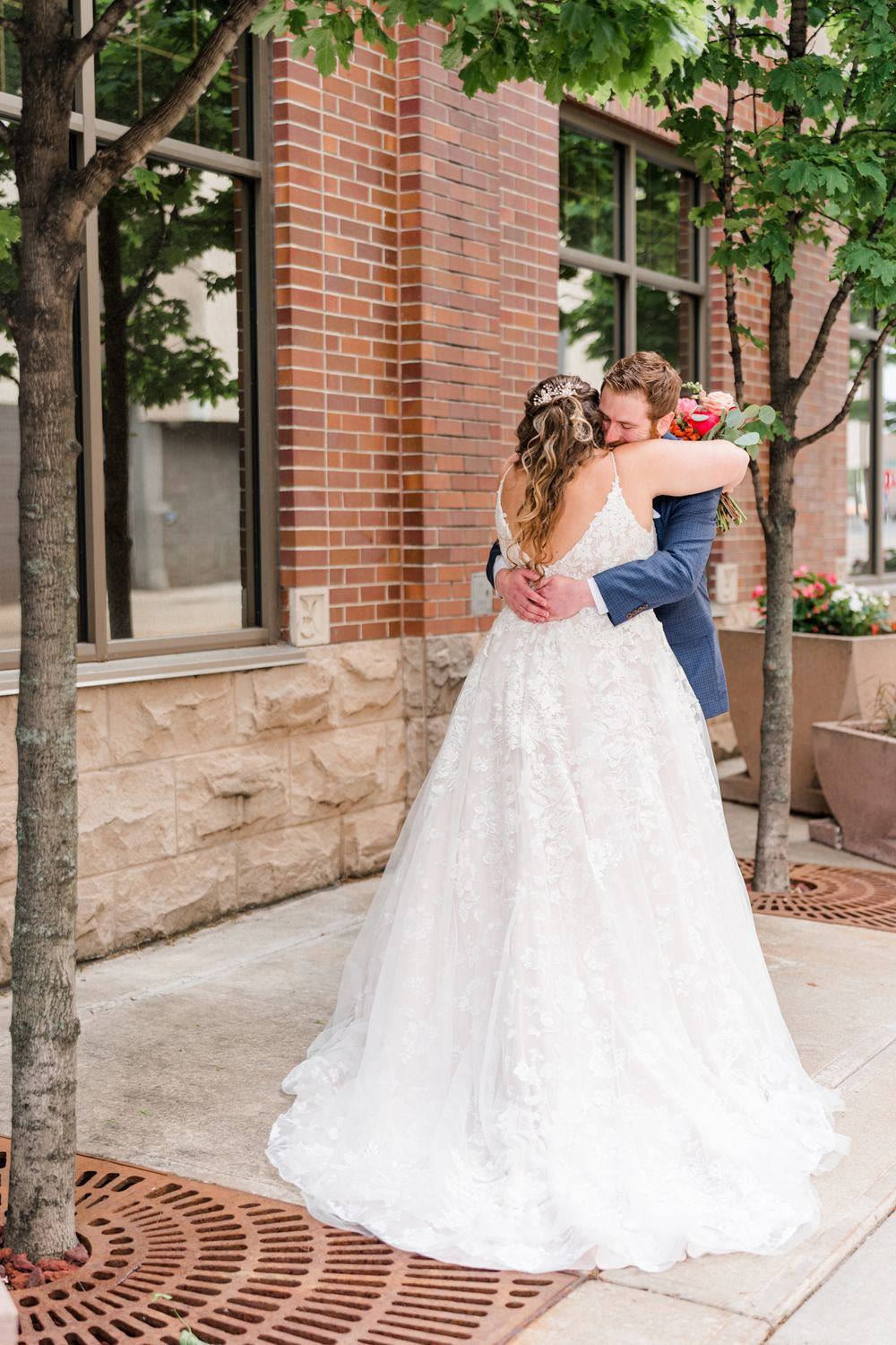 The couple shares a romantic embrace on a tree-lined sidewalk by a brick building.