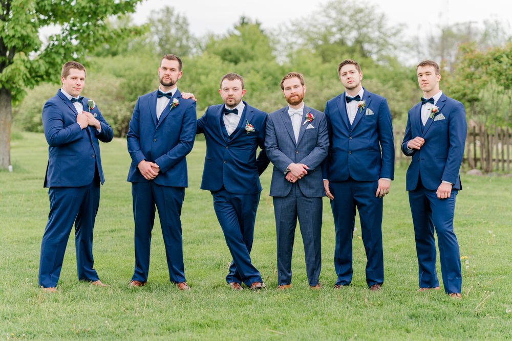 Groomsmen in coordinating navy suits stand together on green lawn during wedding photos.