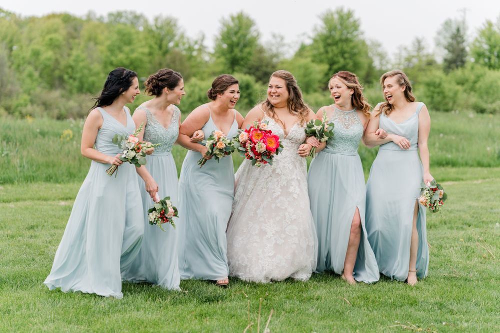 Wedding party in pale blue bridesmaid dresses holding colorful bouquets on grassy hillside.
