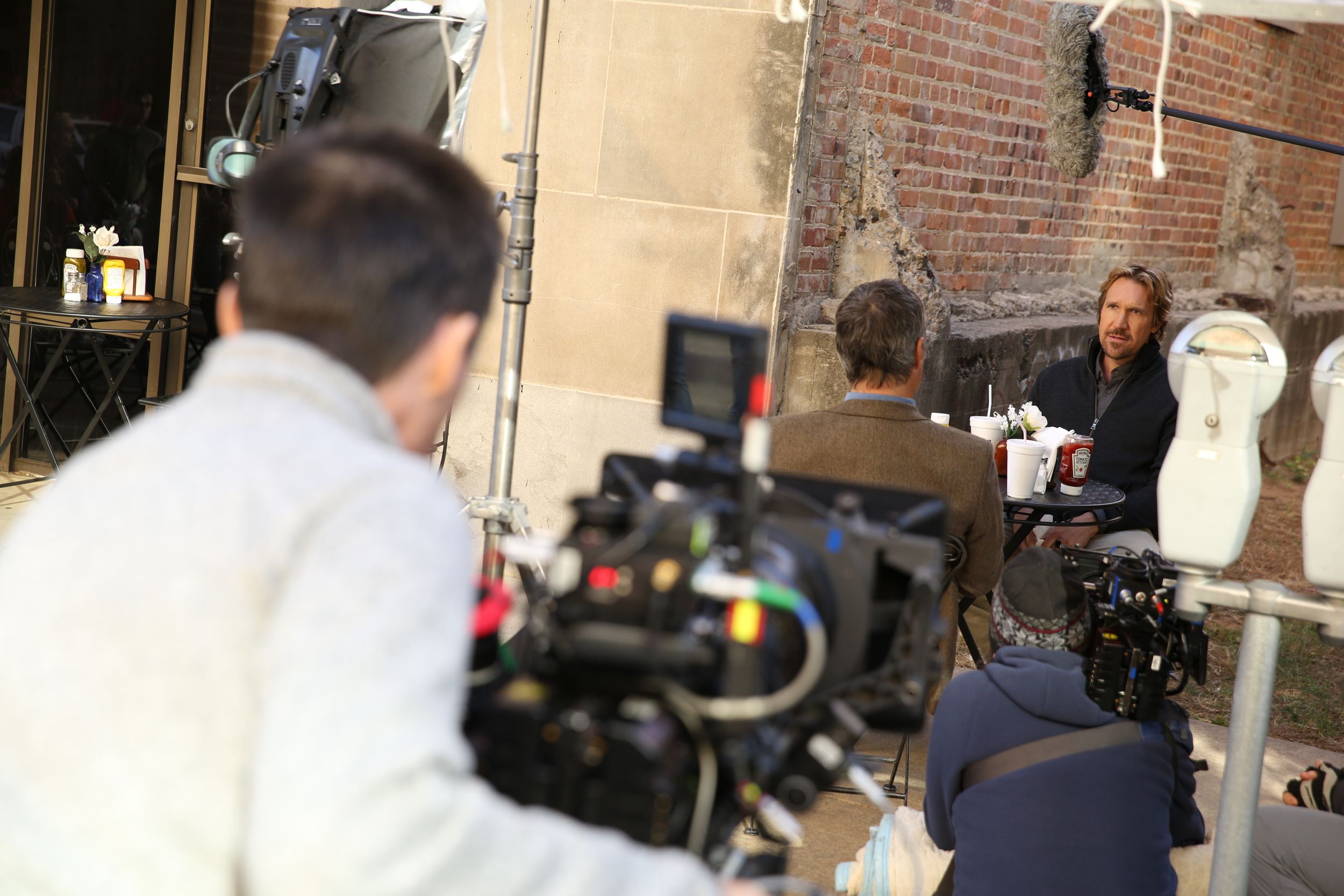 Film crew members setting up equipment against a brick wall backdrop during a production shoot.