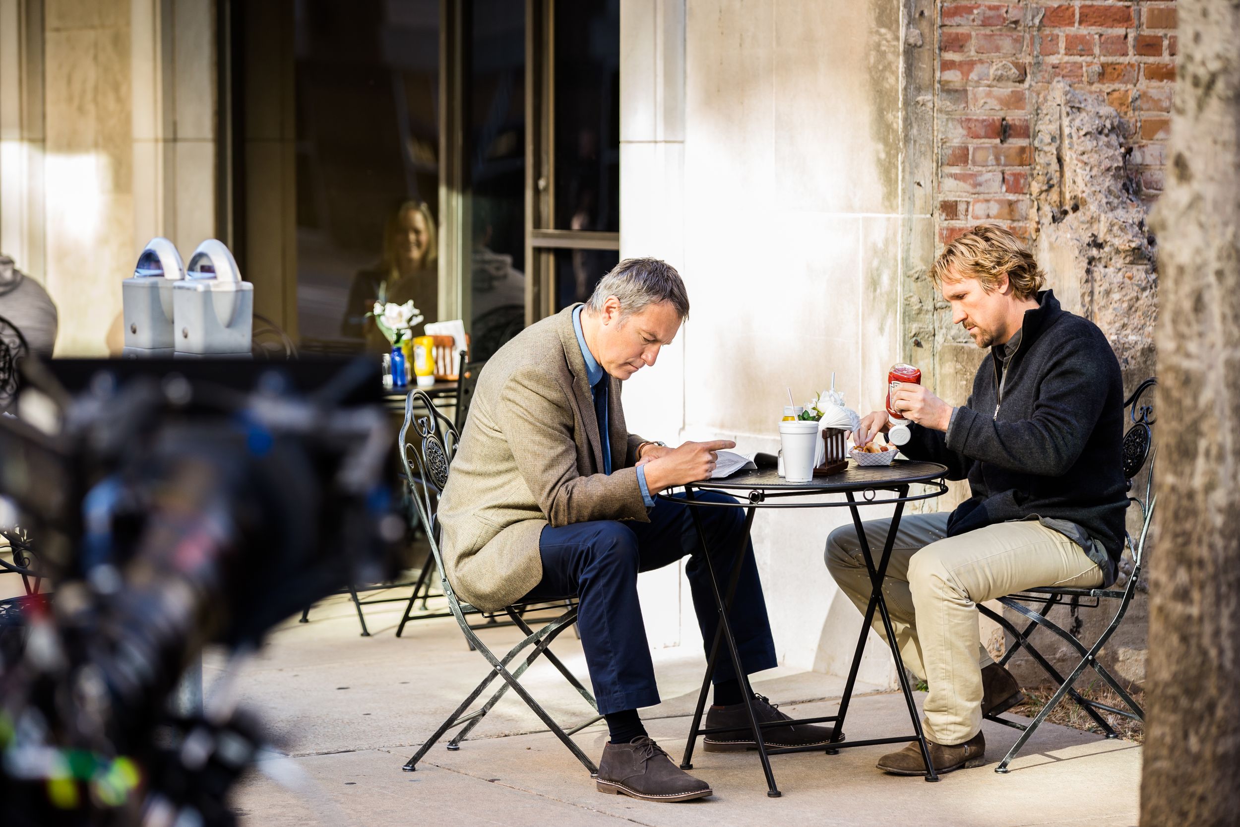 Production team filming a scene with chairs and equipment arranged against an historic brick building exterior.