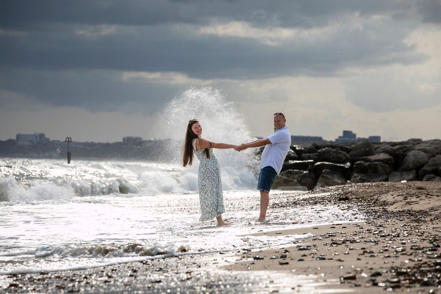 Two people hold hands while walking along a stormy beach with crashing waves.