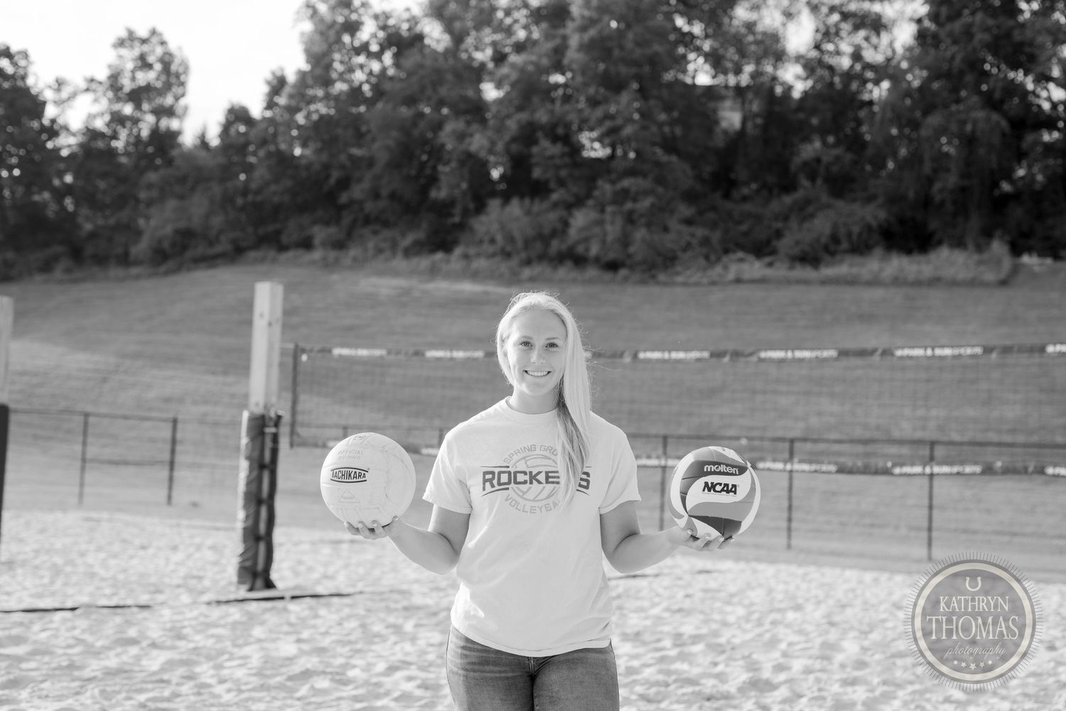 Athlete poses with volleyball equipment on a sand court against a wooded backdrop.