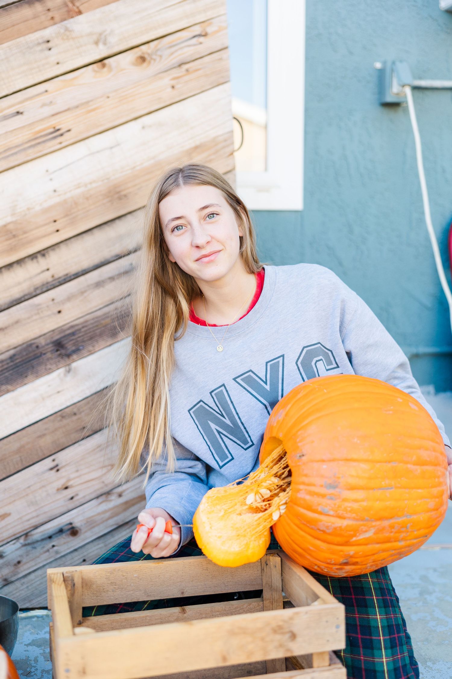 Person in NYC sweatshirt holding small and large pumpkins by wooden wall.