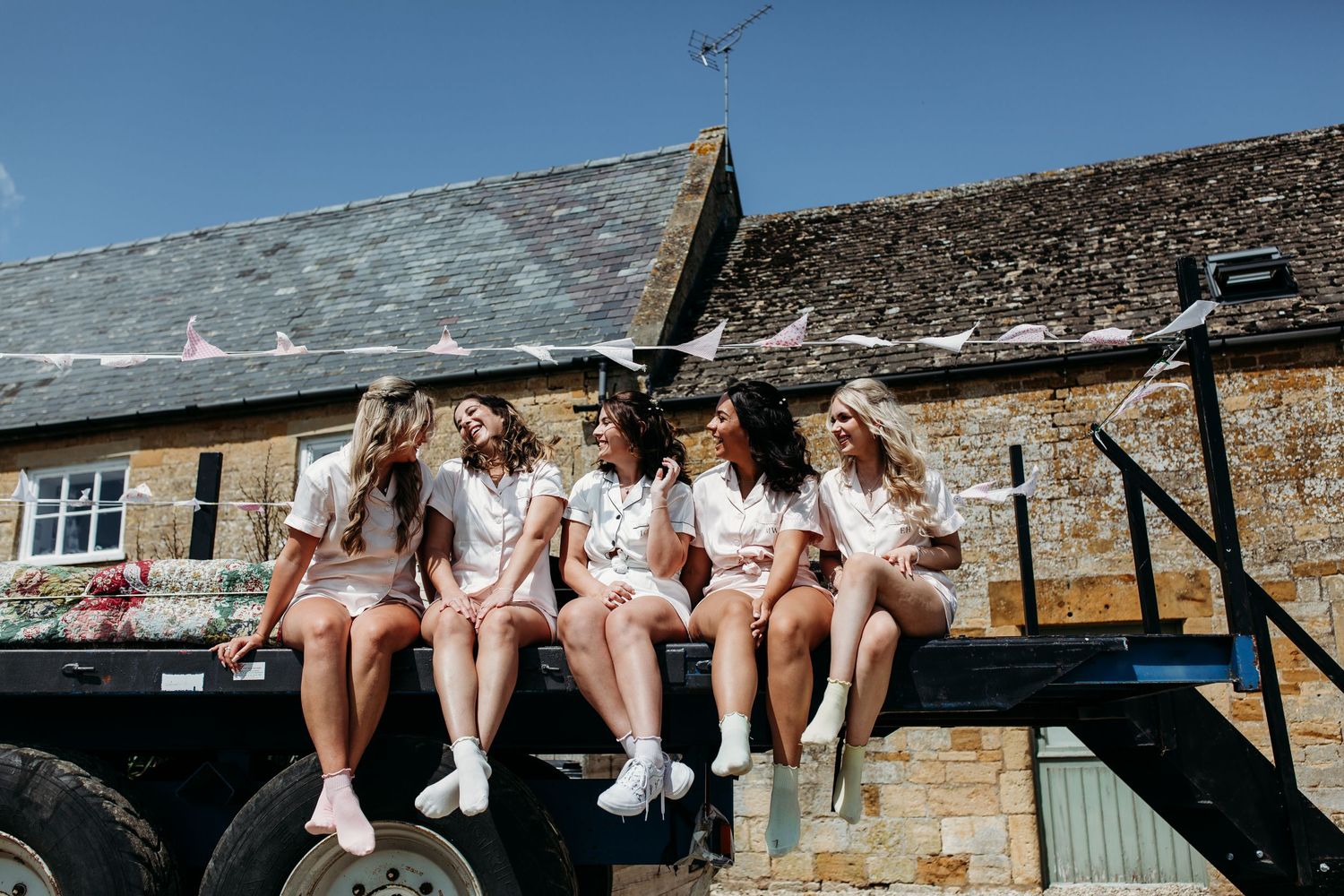 Bridal party in matching white outfits sits casually on rustic farm equipment.