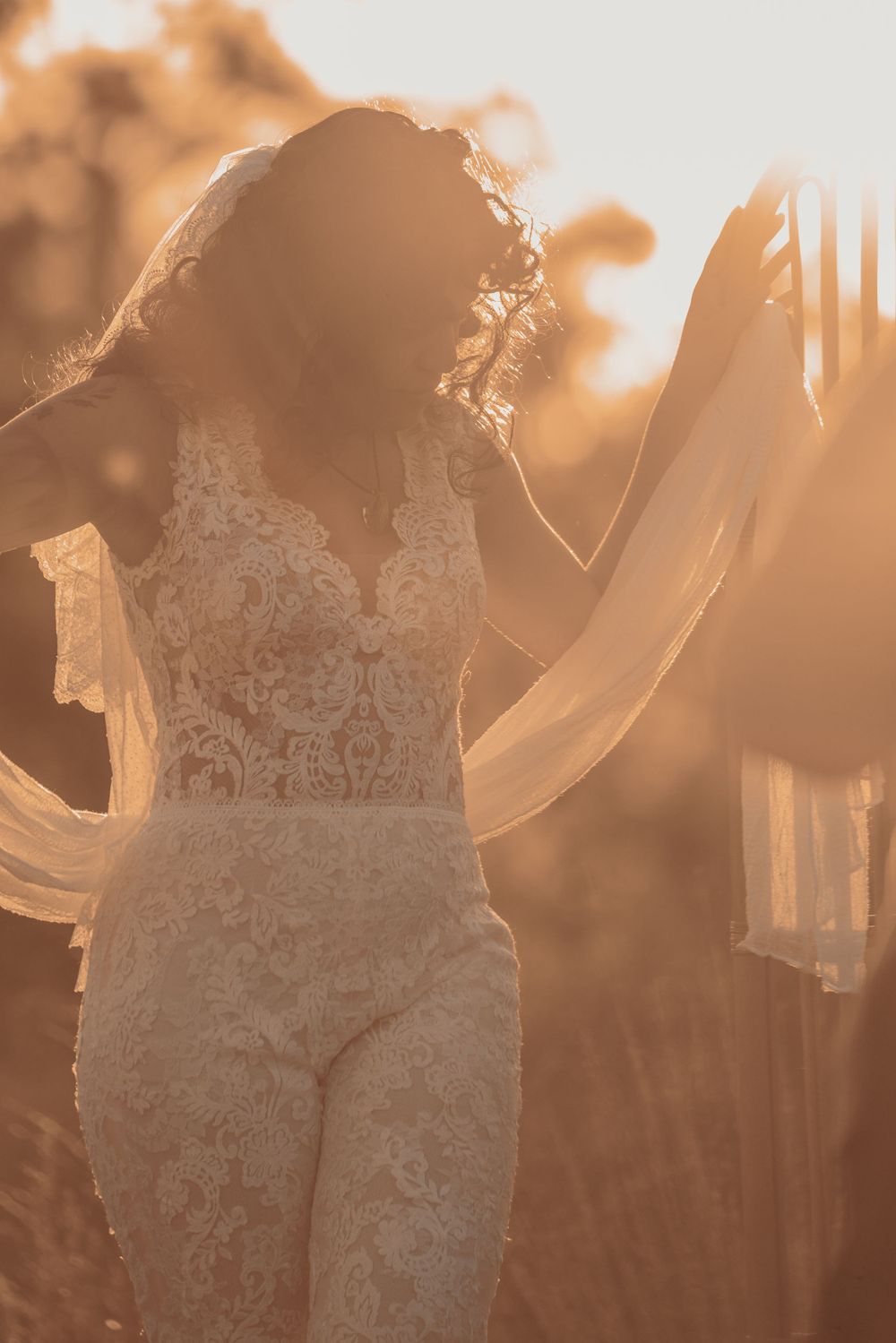 Silhouette of bride in lace jumpsuit with flowing veil at golden hour.