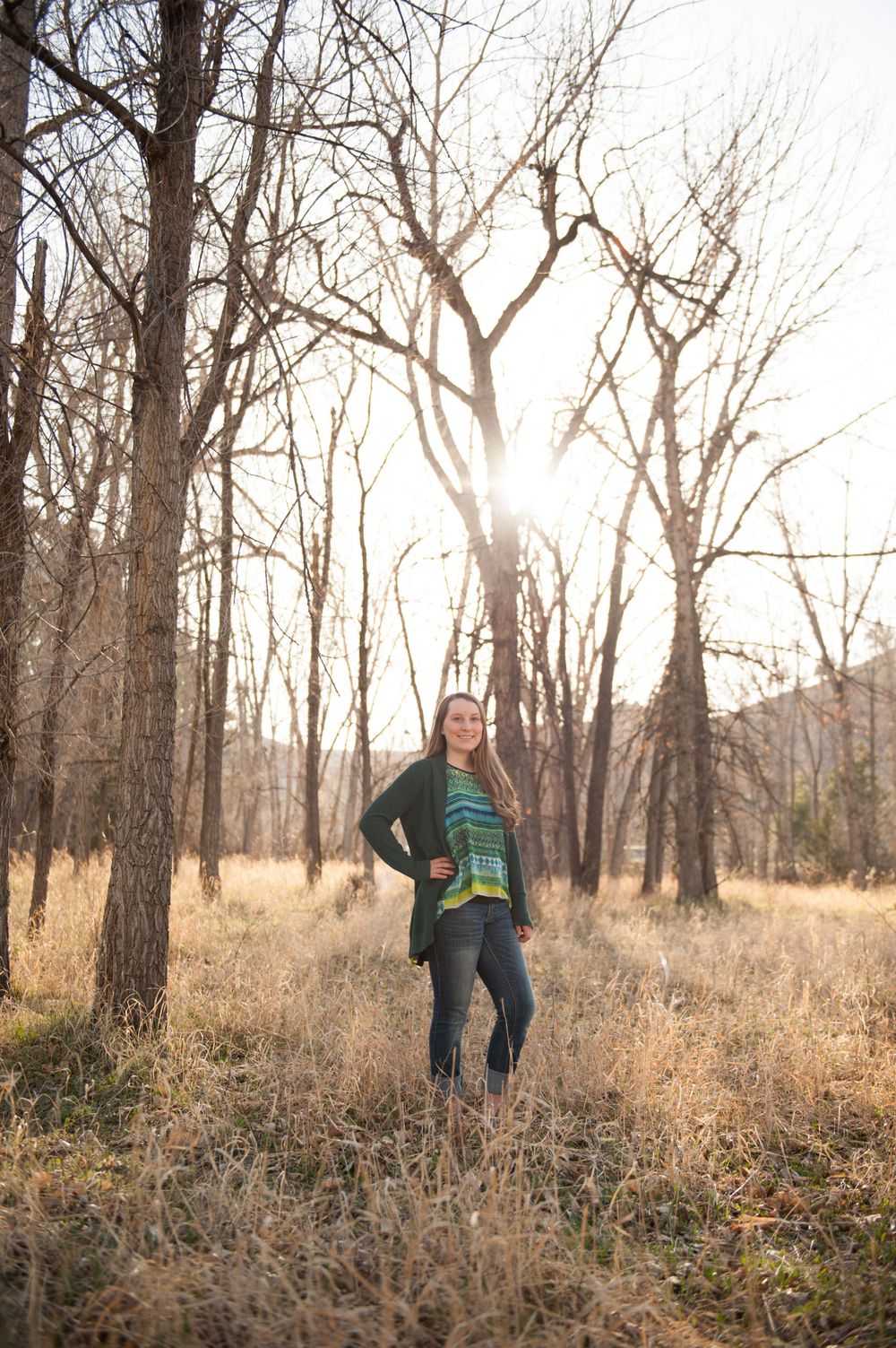 Backlit nature portrait with person in green sweater standing among bare winter trees.