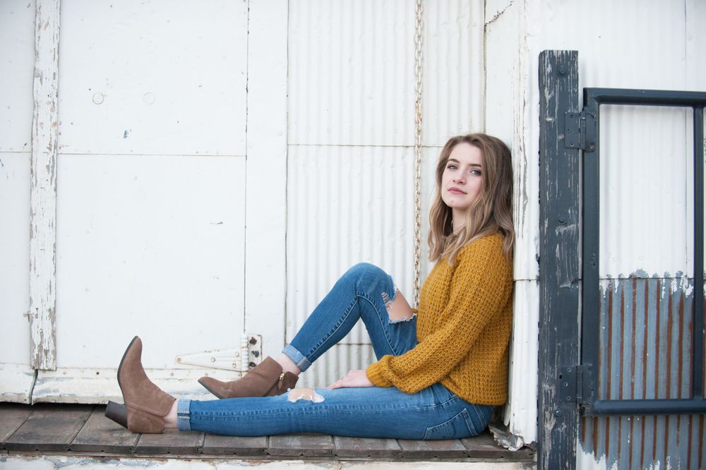 A person in a yellow sweater and jeans sits casually against a white wall wearing brown boots.