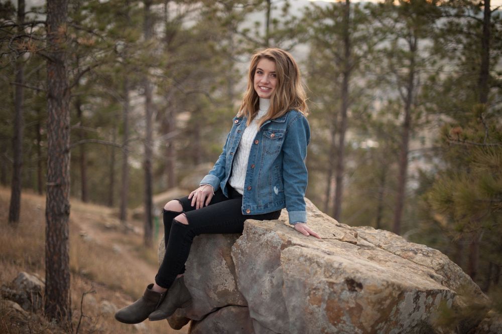 A person in denim jacket sits on a large rock in a mountainous pine forest setting.