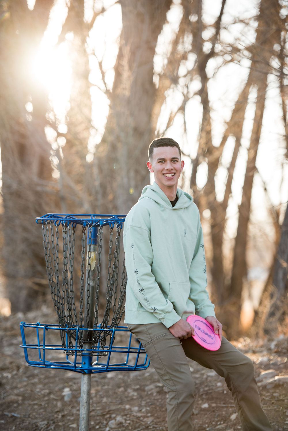 A disc golf player poses with a disc and basket in a sunlit wooded area.