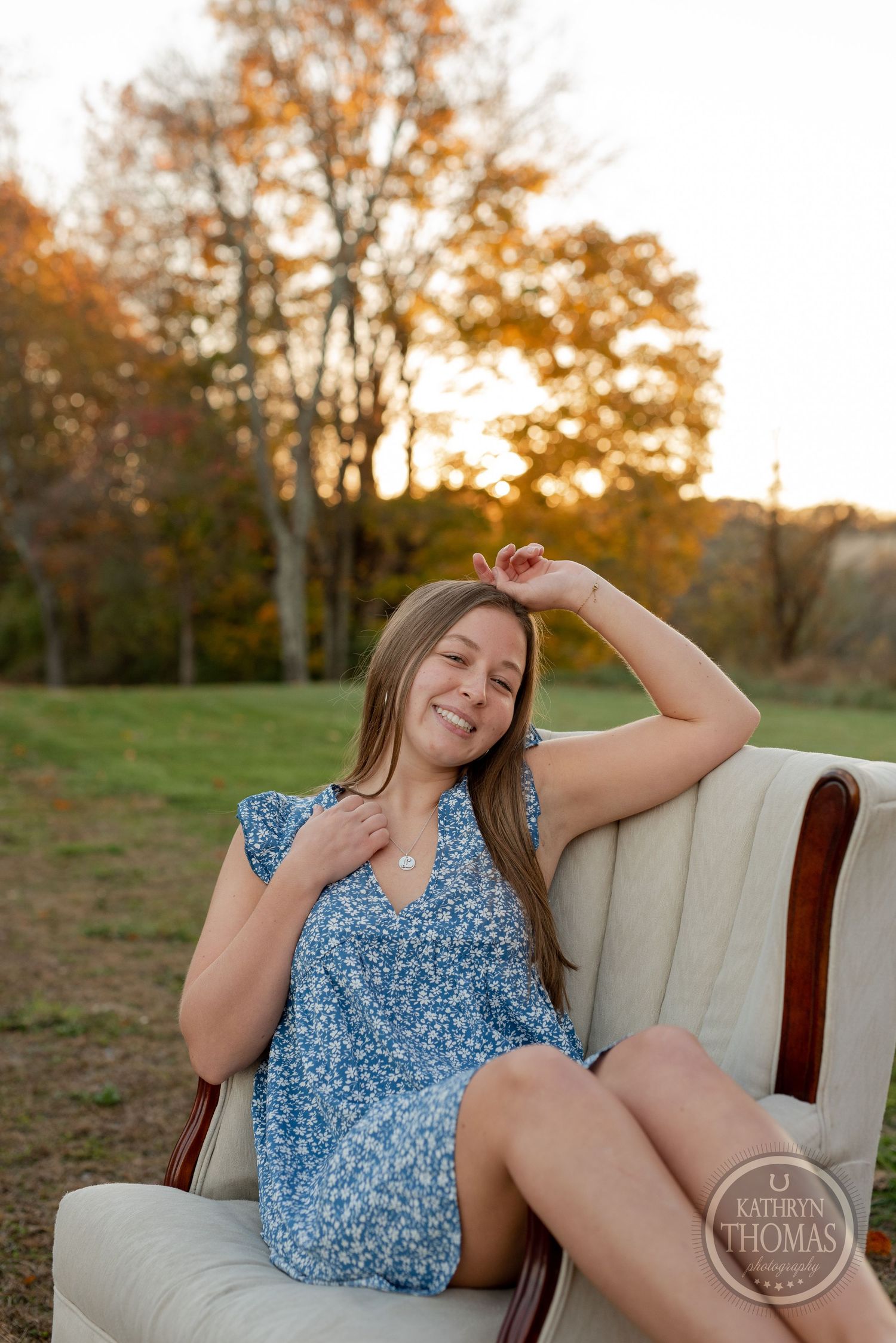 Person in floral blue dress poses playfully in vintage chair during sunset photoshoot.
